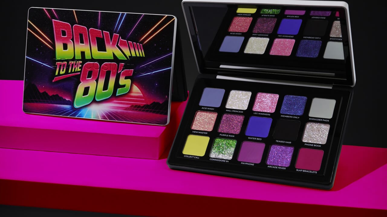 A 16-shade eyeshadow palette set displayed on a magenta platform against a black background. The closed palette on the left features a colorful retro 'Back to the 80's' design, while the open palette on the right displays various glitter and matte shades including pinks, purples, blues, greens, and yellows.
