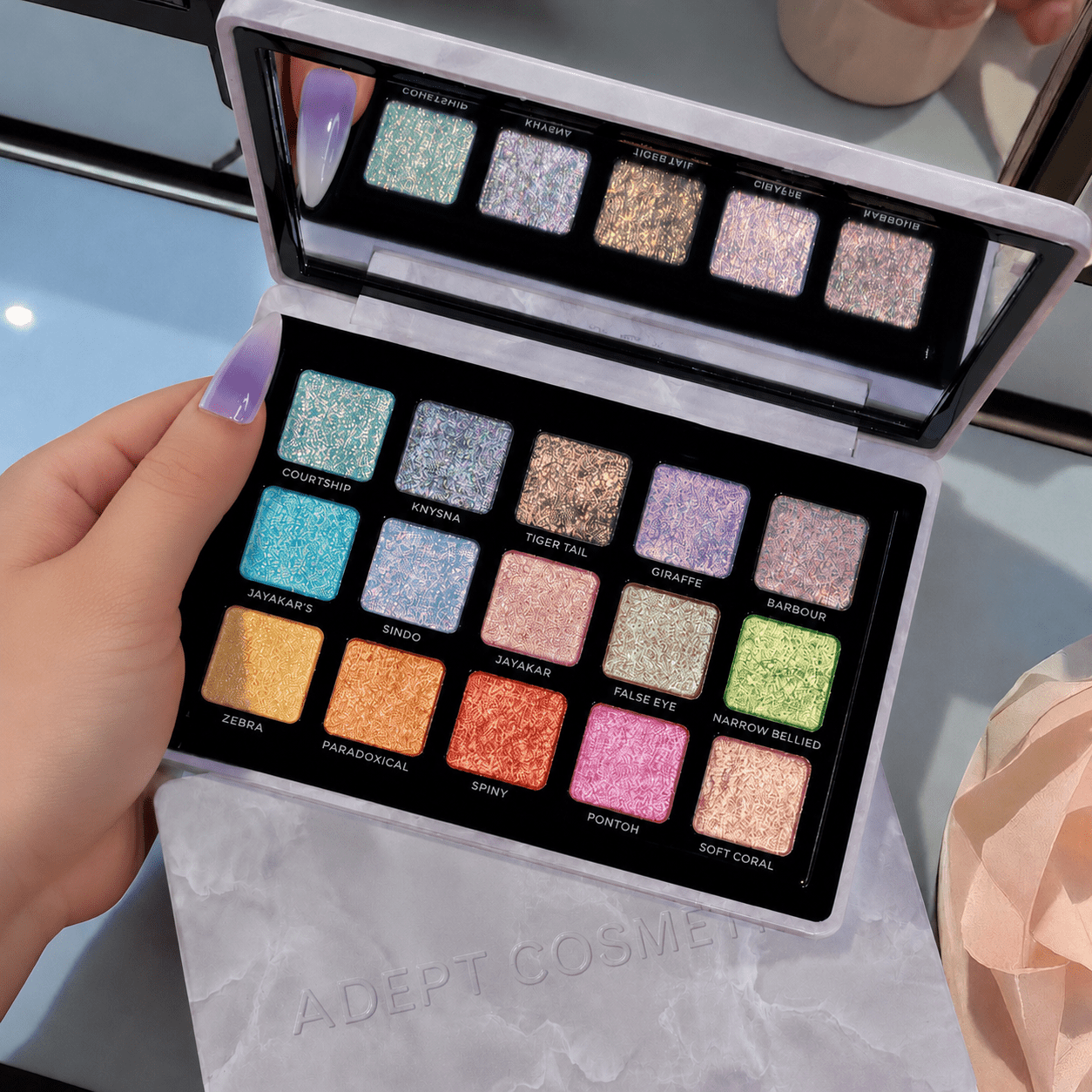 Open makeup palette with glittery eyeshadow shades held by a hand, brand 'Adept Cosmetics' visible.