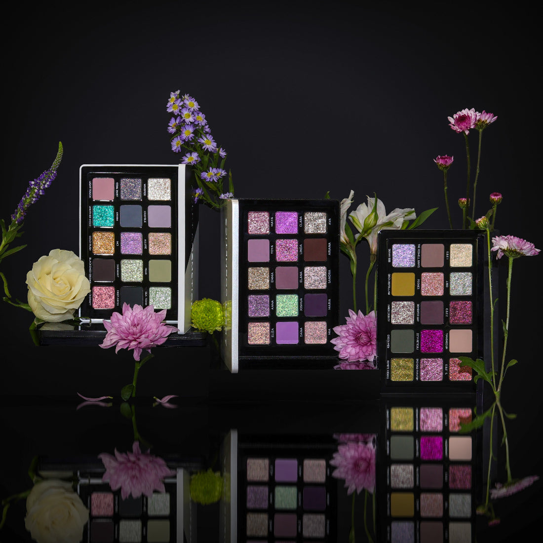 Makeup palettes with colorful shades surrounded by flowers on a reflective surface.