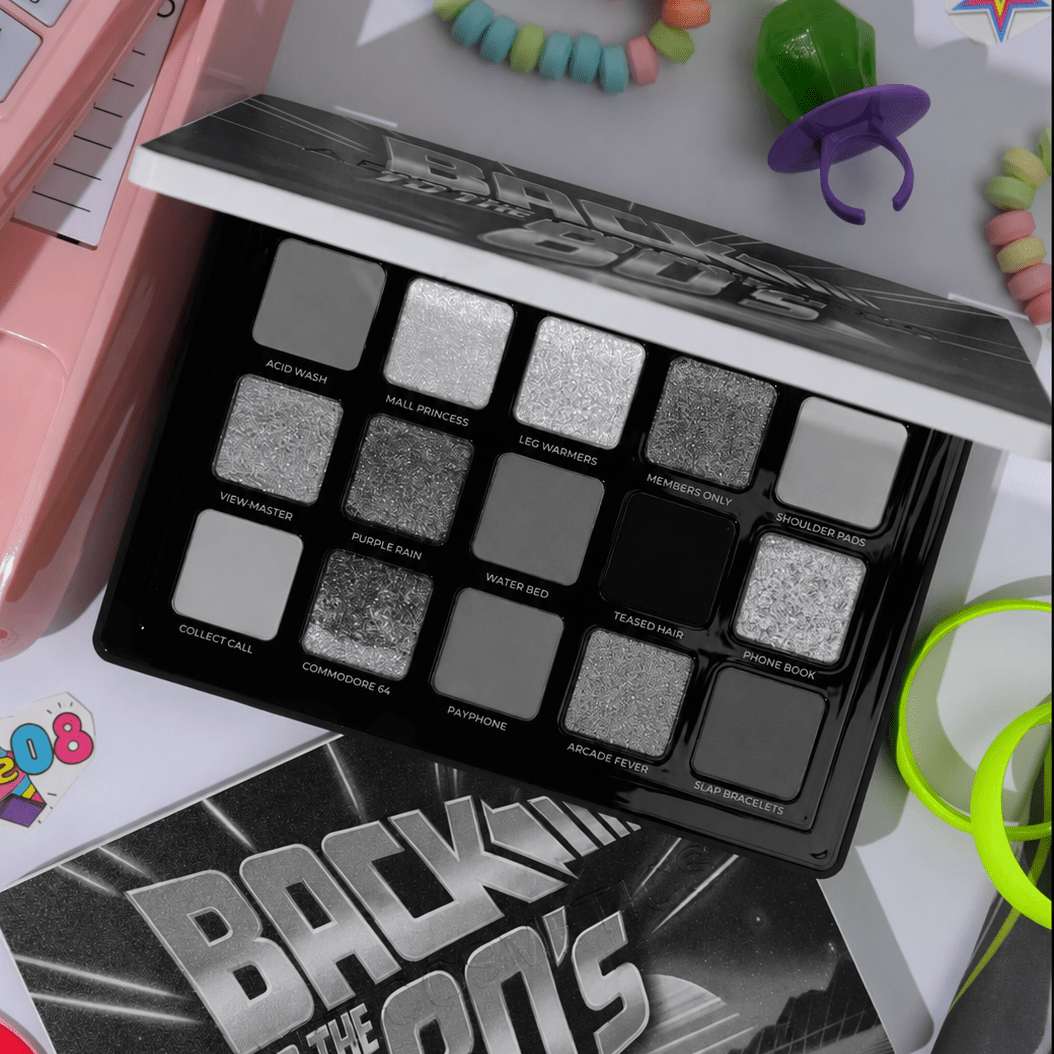 A Back to the 80's themed eyeshadow palette with 12 shades in black, gray, and silver, displayed alongside its matching box and colorful retro-themed props.